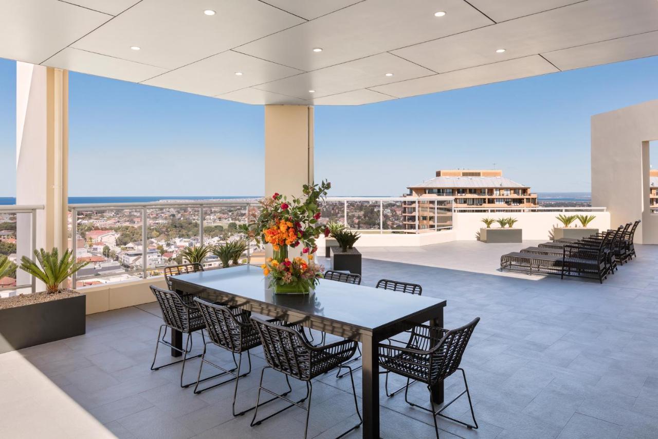 Meriton Suites Bondi Junction Σίδνεϊ Εξωτερικό φωτογραφία The photo shows a stylish outdoor terrace or balcony area, featuring a long dining table surrounded by several black wire chairs. The table has a centerpiece of vibrant flowers. The space is well-lit, with a clear view of the horizon in the backgroun