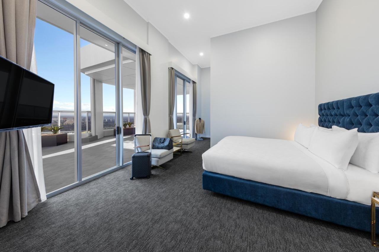 Meriton Suites Bondi Junction Σίδνεϊ Εξωτερικό φωτογραφία The photo shows a modern and spacious hotel room. It features a large bed with a blue headboard, a flat-screen TV mounted on the wall, and a cozy seating area with a chair and small table. The room has large windows that offer natural light and a vie