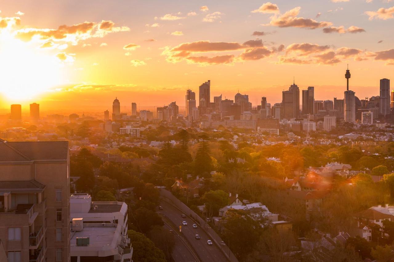 Meriton Suites Bondi Junction Σίδνεϊ Εξωτερικό φωτογραφία The photo shows a panoramic view of a city skyline at sunset. The sun is low on the horizon, casting a warm golden light across the buildings. Tall skyscrapers are silhouetted against the colorful sky, which features hues of orange, pink, and yellow,