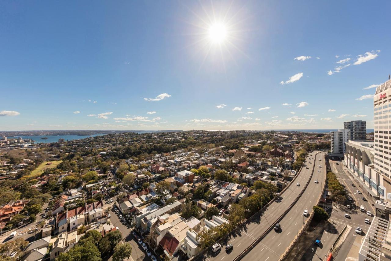 Meriton Suites Bondi Junction Σίδνεϊ Εξωτερικό φωτογραφία The photo shows a panoramic view of a city landscape under a clear blue sky. In the foreground, there is a road with vehicles traveling along it, bordered by trees and buildings. In the background, you can see a residential area with various houses, 