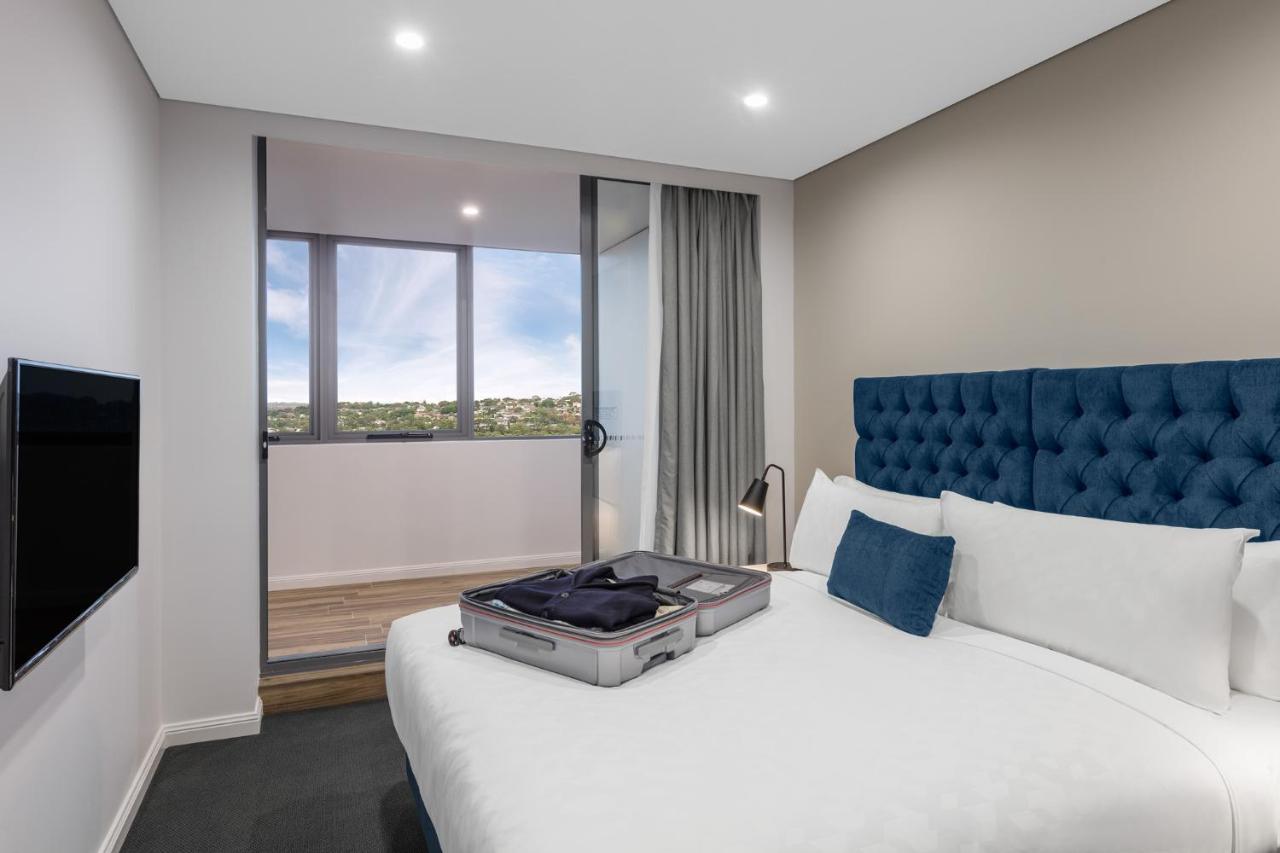 Meriton Suites Bondi Junction Σίδνεϊ Εξωτερικό φωτογραφία The photo shows a modern hotel room featuring a double bed with a tufted blue headboard. There is a neatly arranged suitcase placed open on the bed, indicating that someone may be unpacking. The room has light-colored walls and a window allowing natu