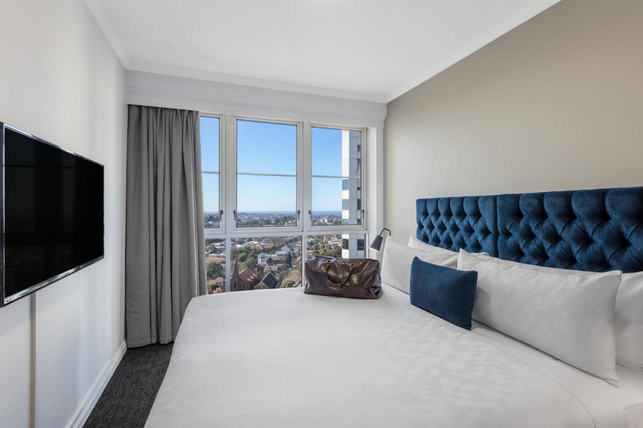 Meriton Suites Bondi Junction Σίδνεϊ Εξωτερικό φωτογραφία The photo shows a modern hotel room featuring a large bed with a tufted blue headboard. Beside the bed, there is a stylish bag placed on the bed. The room has large windows that provide a view of the outside, allowing natural light to fill the space.