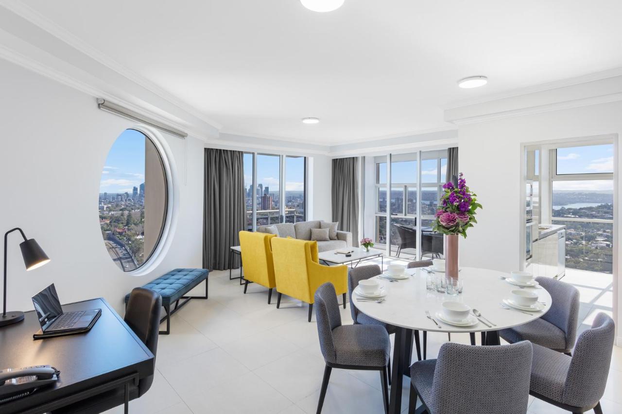Meriton Suites Bondi Junction Σίδνεϊ Εξωτερικό φωτογραφία The photo shows a modern and bright interior space, likely a living room or dining area. It features large windows that offer a panoramic view of the city skyline. The furnishings include a round dining table set for several people, with plates and g