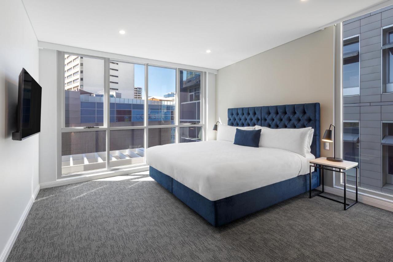 Meriton Suites Bondi Junction Σίδνεϊ Εξωτερικό φωτογραφία The image shows a modern hotel room with a large, comfortable bed featuring a blue upholstered headboard. The room has large windows that allow natural light to enter, providing a good view of the surrounding buildings. There is a small bedside table