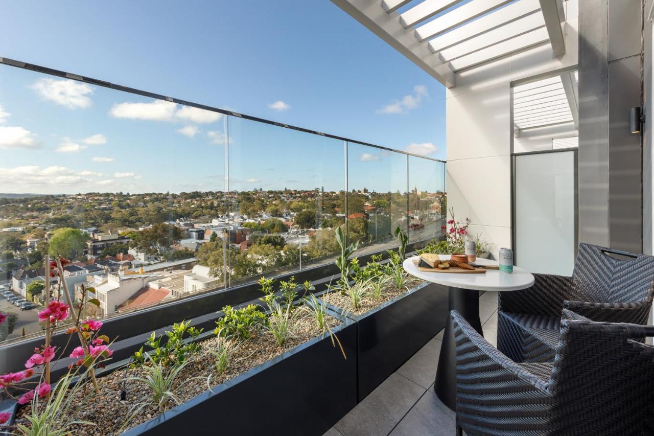 Meriton Suites Bondi Junction Σίδνεϊ Εξωτερικό φωτογραφία The photo shows a modern balcony with a glass railing, enhancing the view of the surrounding area, which includes various rooftops and greenery in the distance. There is a small round table set with a decorative floral arrangement and some items, lik