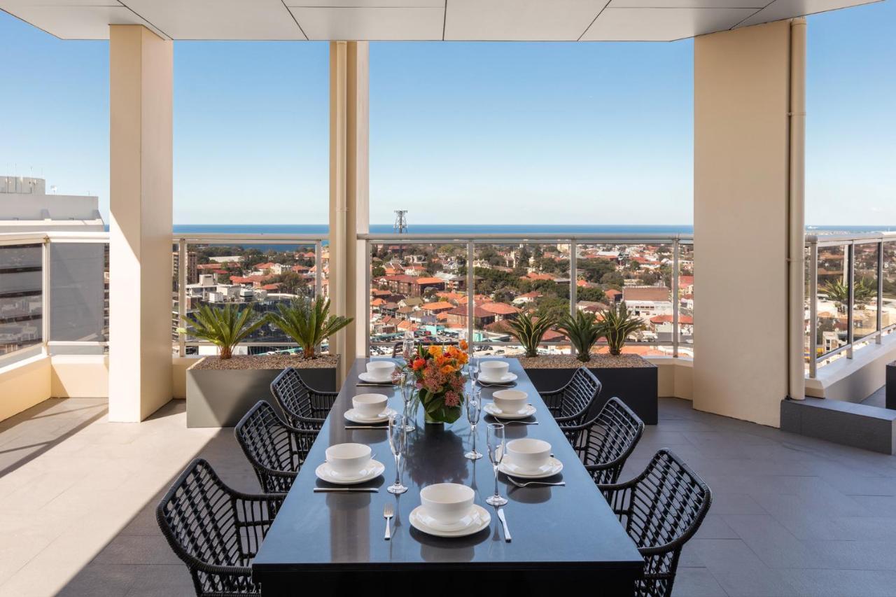 Meriton Suites Bondi Junction Σίδνεϊ Εξωτερικό φωτογραφία The photo shows a modern outdoor dining area situated on a balcony or terrace with a stunning view. There is a long rectangular table set with plates, glasses, and cutlery, ready for a meal. The table is surrounded by sleek, black wicker chairs. In t