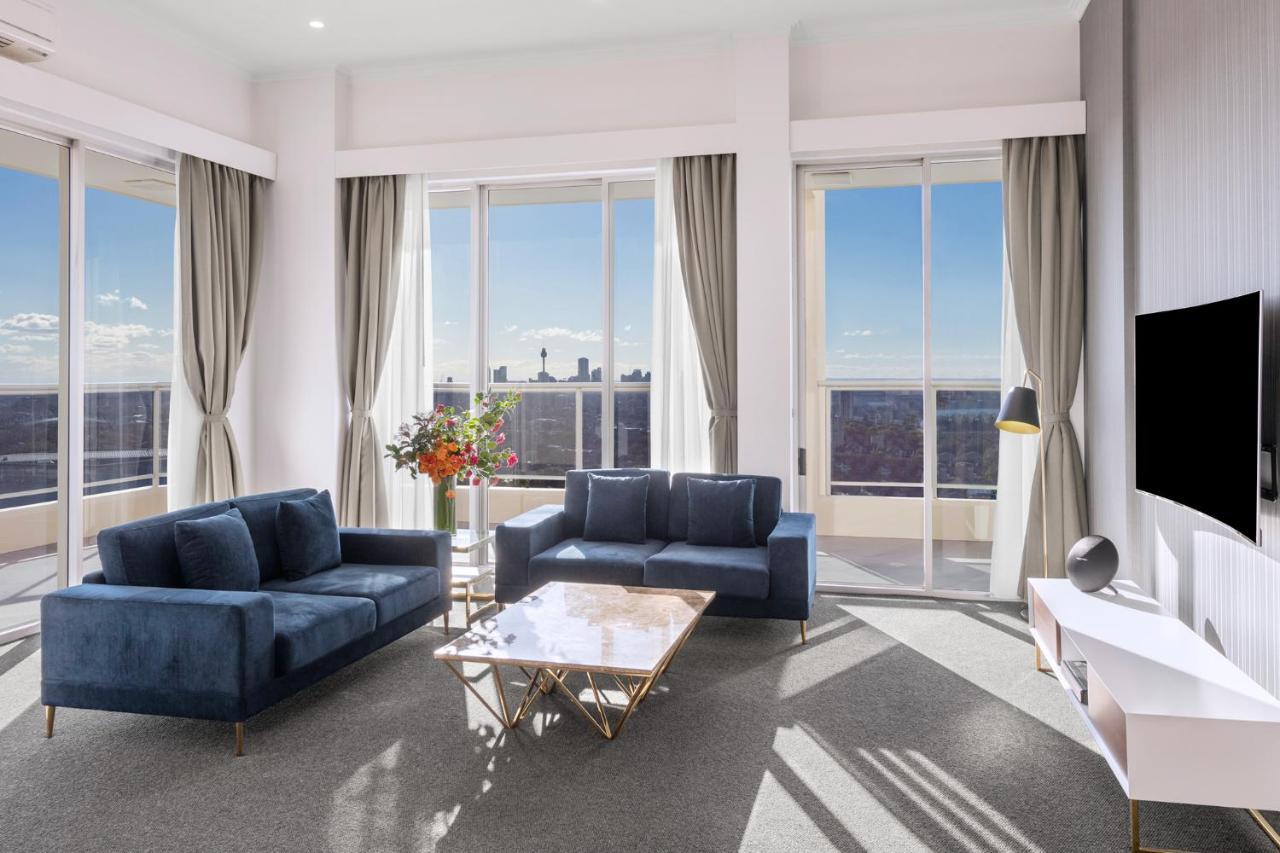 Meriton Suites Bondi Junction Σίδνεϊ Εξωτερικό φωτογραφία The photo shows a modern living room with large windows that allow natural light to fill the space. There are two blue sofas arranged around a coffee table with a glass top and gold legs. A floral arrangement adds a touch of color, placed on a nearby