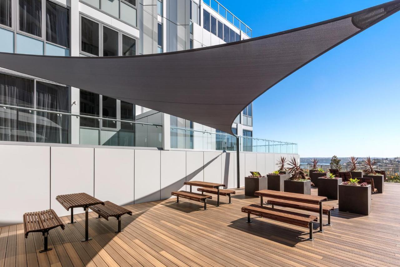 Meriton Suites Bondi Junction Σίδνεϊ Εξωτερικό φωτογραφία The photo shows a modern outdoor terrace area on a building. It features several wooden benches arranged on a wooden deck. There are large planters with greenery, adding some decorative elements. A large triangular gray shade sail is positioned above