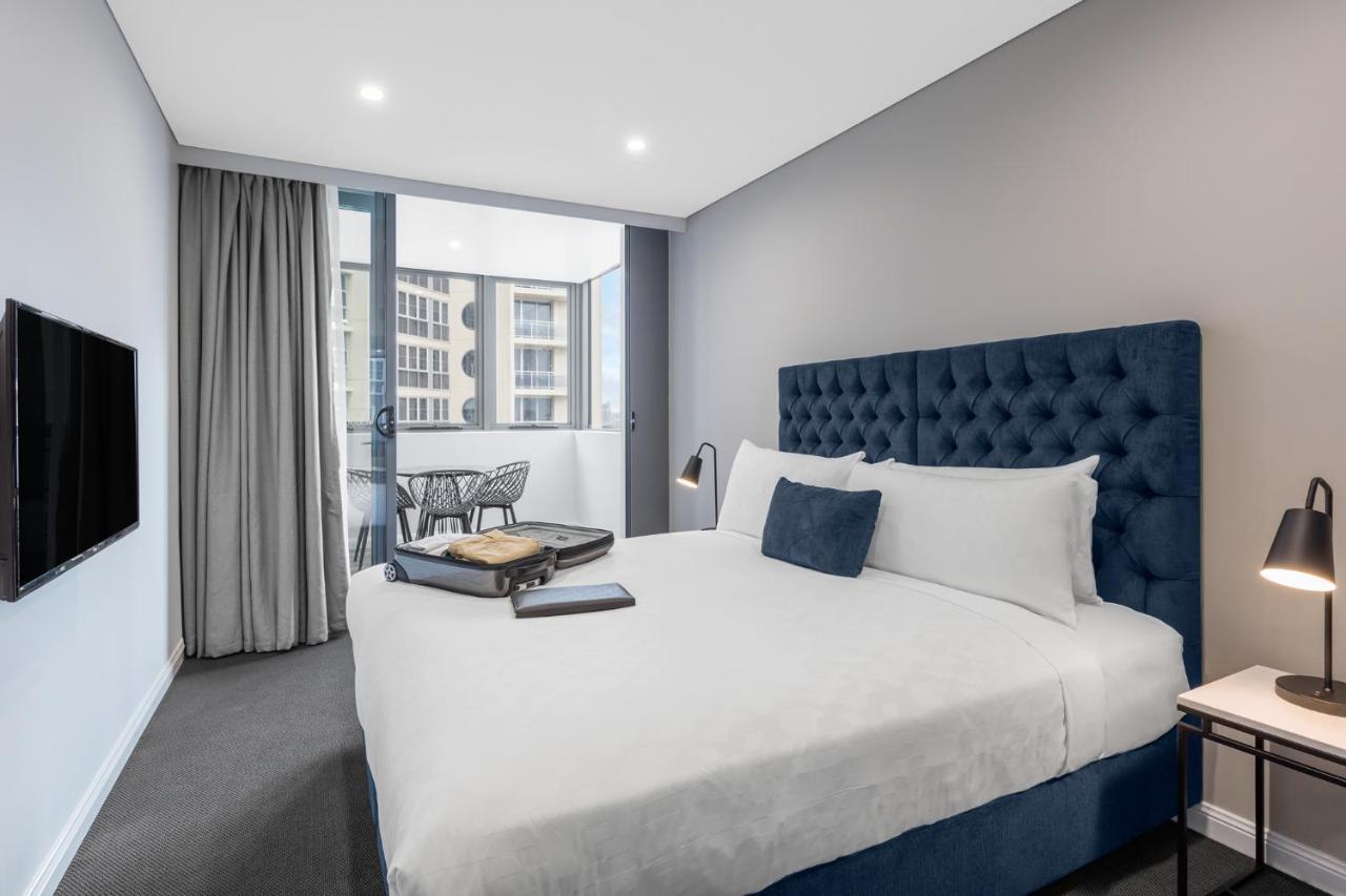 Meriton Suites Bondi Junction Σίδνεϊ Εξωτερικό φωτογραφία The photo shows a modern hotel room featuring a large bed with a tufted blue headboard. There are minimalistic bedside tables with lamps on either side of the bed. A flat-screen TV is mounted on the wall across from the bed. To one side, there are fl