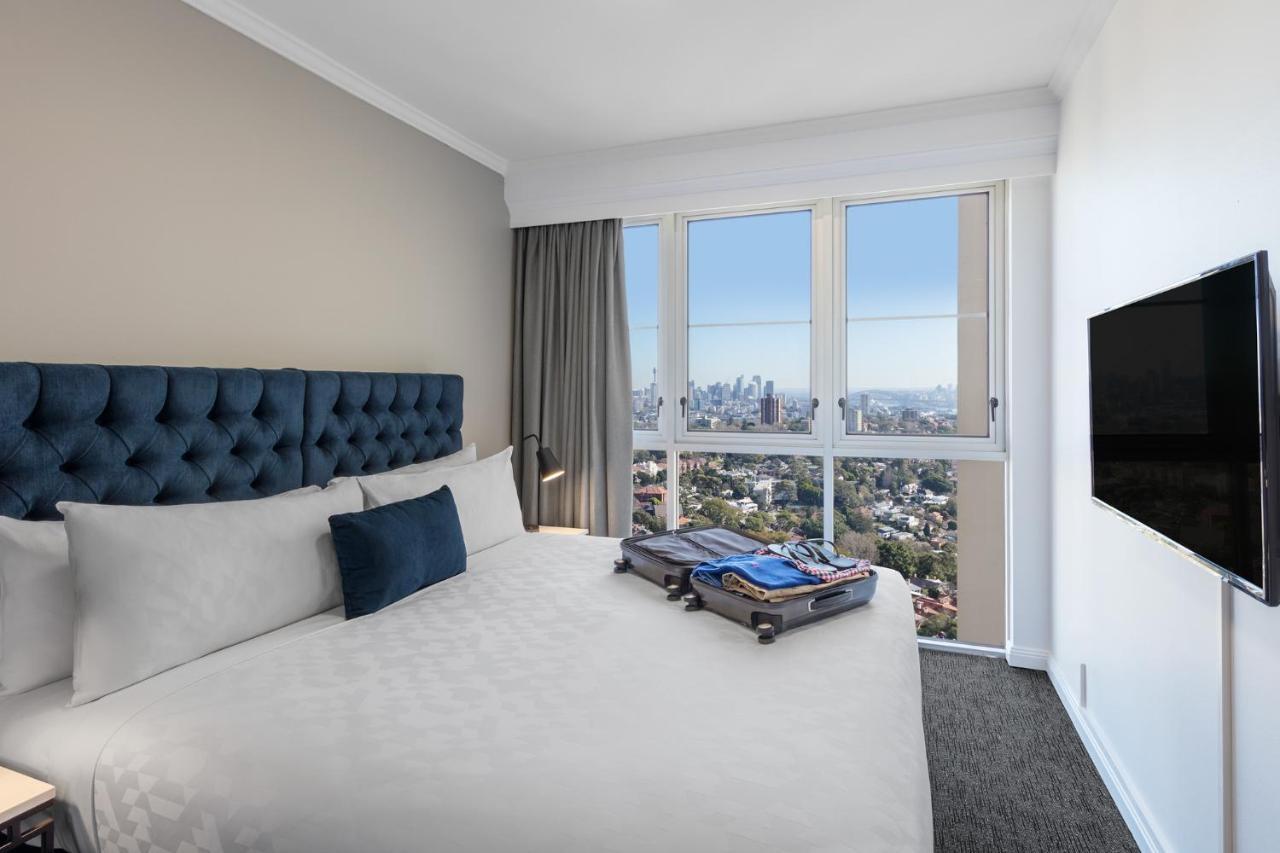 Meriton Suites Bondi Junction Σίδνεϊ Εξωτερικό φωτογραφία The photo shows a modern hotel bedroom featuring a king-sized bed with a blue tufted headboard. The bed is neatly made with white linens and a decorative blue pillow. There is a large window that offers a view of the city skyline, allowing natural li