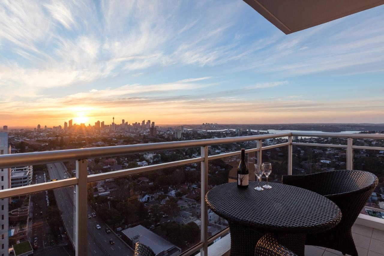 Meriton Suites Bondi Junction Σίδνεϊ Εξωτερικό φωτογραφία The image shows a scenic view from a balcony or terrace overlooking a cityscape during sunset. In the foreground, there is a small round table with two glasses and a bottle of wine, suggesting a romantic or relaxing setting. The sky is filled with vi
