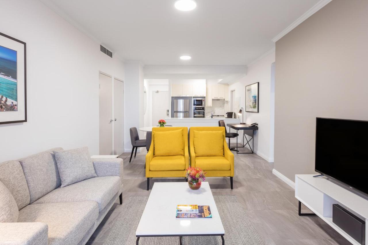 Meriton Suites Bondi Junction Σίδνεϊ Εξωτερικό φωτογραφία The photo shows a modern living space with a light and airy design. In the foreground, there are two bright yellow armchairs flanking a white coffee table, which has a small vase of flowers and a magazine on it. To the left, there is a light gray sof