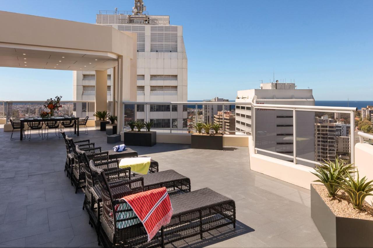 Meriton Suites Bondi Junction Σίδνεϊ Εξωτερικό φωτογραφία The photo shows a rooftop terrace with a modern design. There are several outdoor seating arrangements featuring black wicker lounge chairs. Some of the chairs have colorful blankets draped over them. The terrace is surrounded by a railing and has po