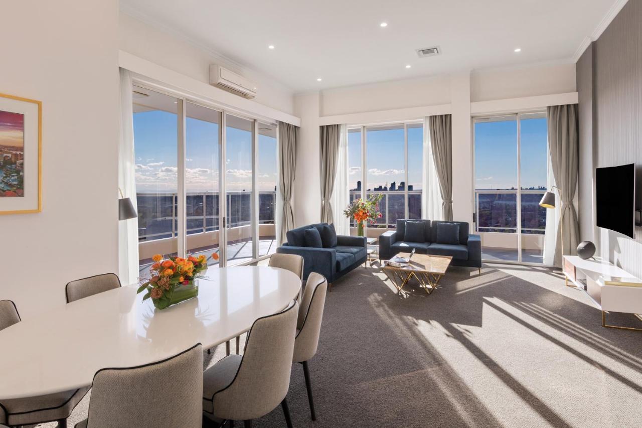 Meriton Suites Bondi Junction Σίδνεϊ Εξωτερικό φωτογραφία The photo shows a modern and stylish living space with large windows allowing natural light to fill the room. In the foreground, there is a round dining table with several chairs around it, adorned with a floral centerpiece. The seating area features