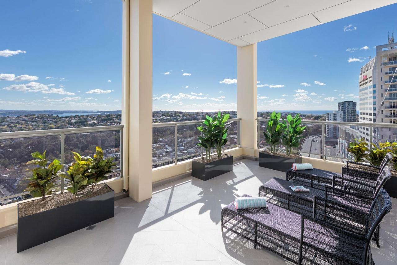 Meriton Suites Bondi Junction Σίδνεϊ Εξωτερικό φωτογραφία The photo shows a modern balcony or terrace area with a stunning view. There are several planters with green plants placed along the edge. The seating area features lounge chairs in a contemporary design, likely made of woven materials. The space app