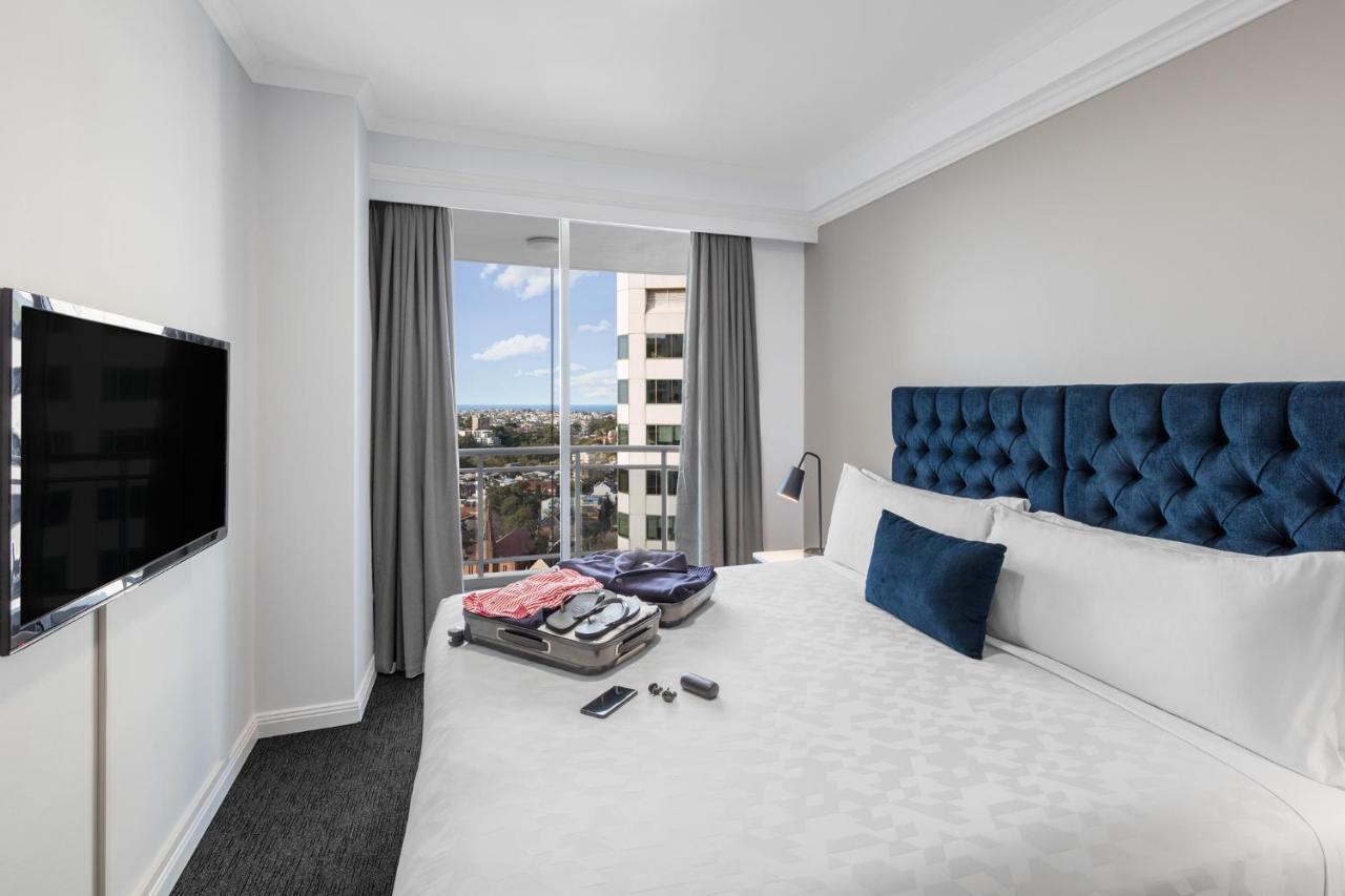 Meriton Suites Bondi Junction Σίδνεϊ Εξωτερικό φωτογραφία The photo shows a modern hotel room featuring a neatly made bed with a blue tufted headboard and a white duvet. There's a small decorative pillow on the bed, and a piece of luggage is placed at the foot with clothes partially unpacked. A television i