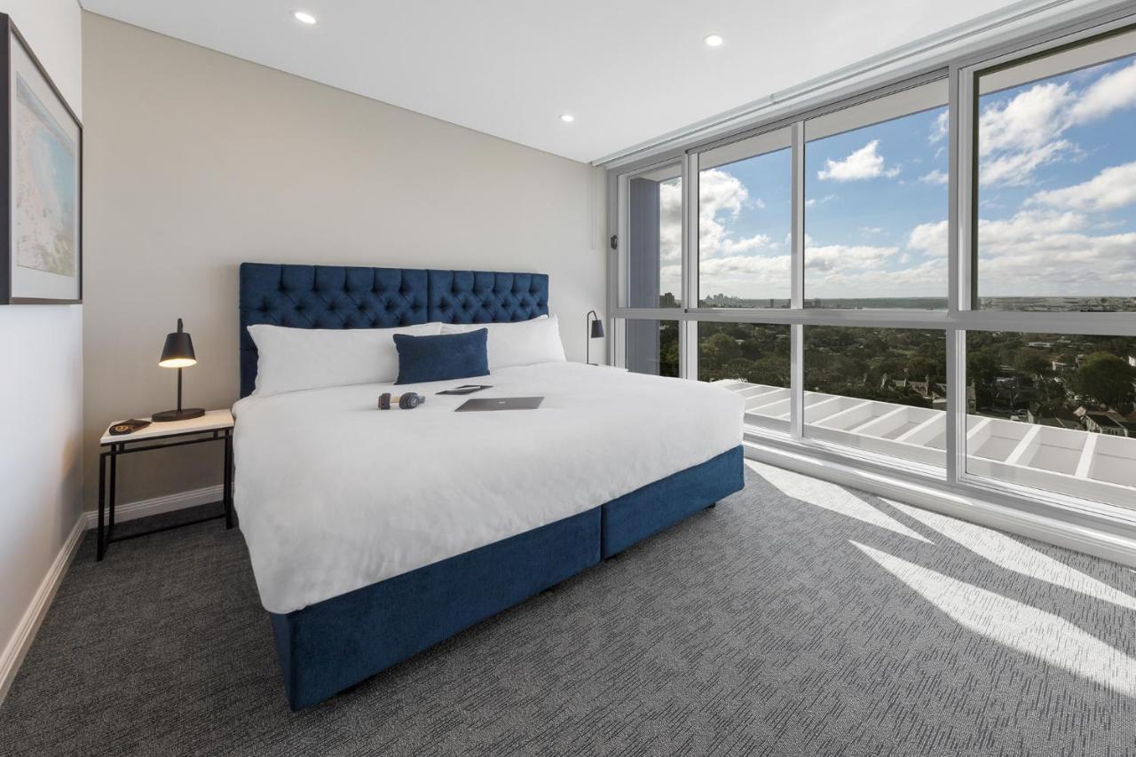 Meriton Suites Bondi Junction Σίδνεϊ Εξωτερικό φωτογραφία The photo shows a modern and stylish hotel room. It features a comfortable queen-size bed with a blue tufted headboard. The bed is neatly made with white linens and pillows. Next to the bed, there is a small bedside table with a desk lamp. 

On the f