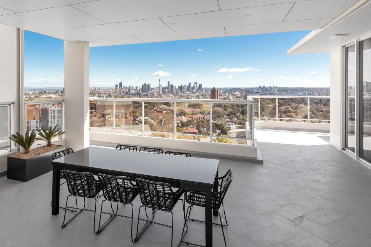 Meriton Suites Bondi Junction Σίδνεϊ Εξωτερικό φωτογραφία The photo shows a modern outdoor balcony or terrace space with a dining table and chairs. The table is black and has a minimalist design, surrounded by stylish chairs. In the background, there is a stunning view of a city skyline under a clear blue s