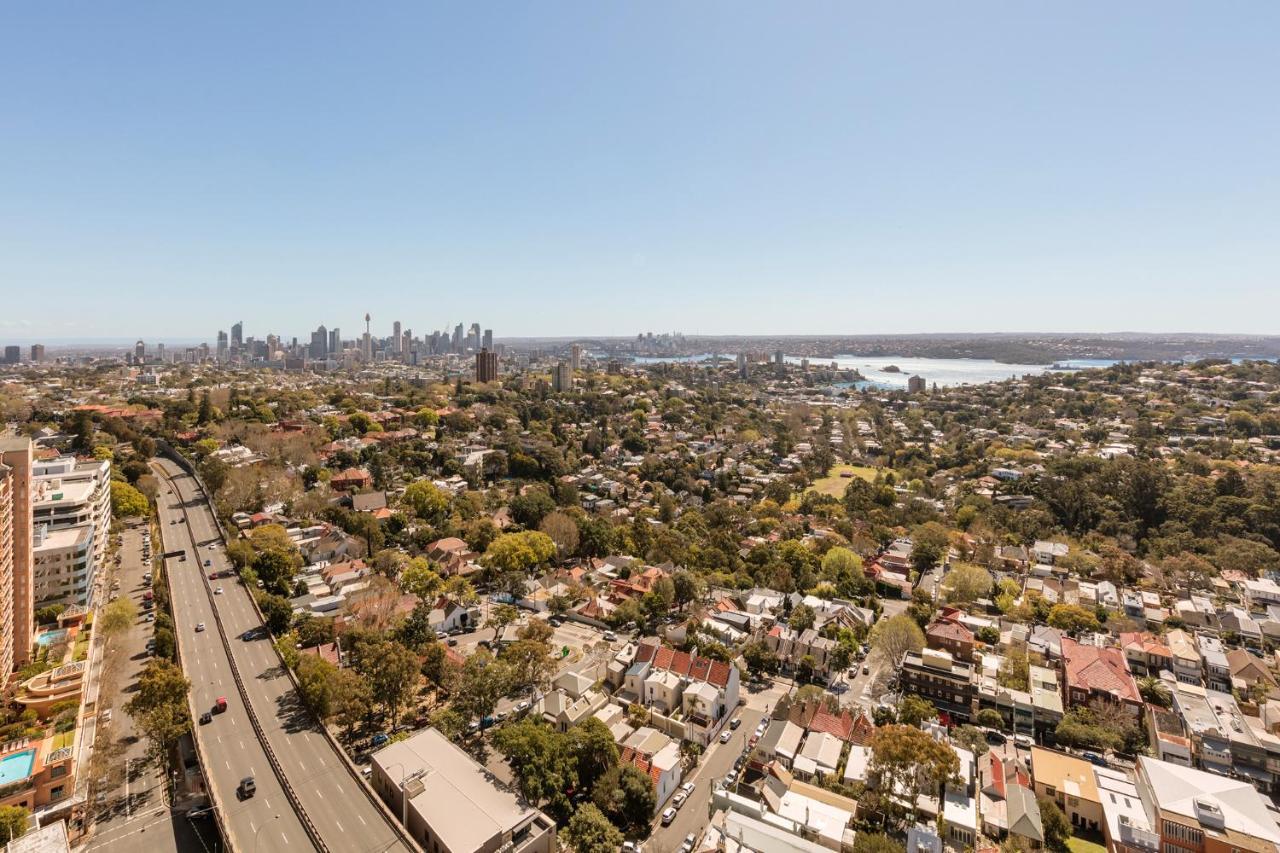 Meriton Suites Bondi Junction Σίδνεϊ Εξωτερικό φωτογραφία The photo captures a panoramic view of a vibrant urban landscape. In the foreground, there are residential areas with various houses and greenery, showcasing a suburban environment. In the background, the skyline of a city can be seen, featuring tall