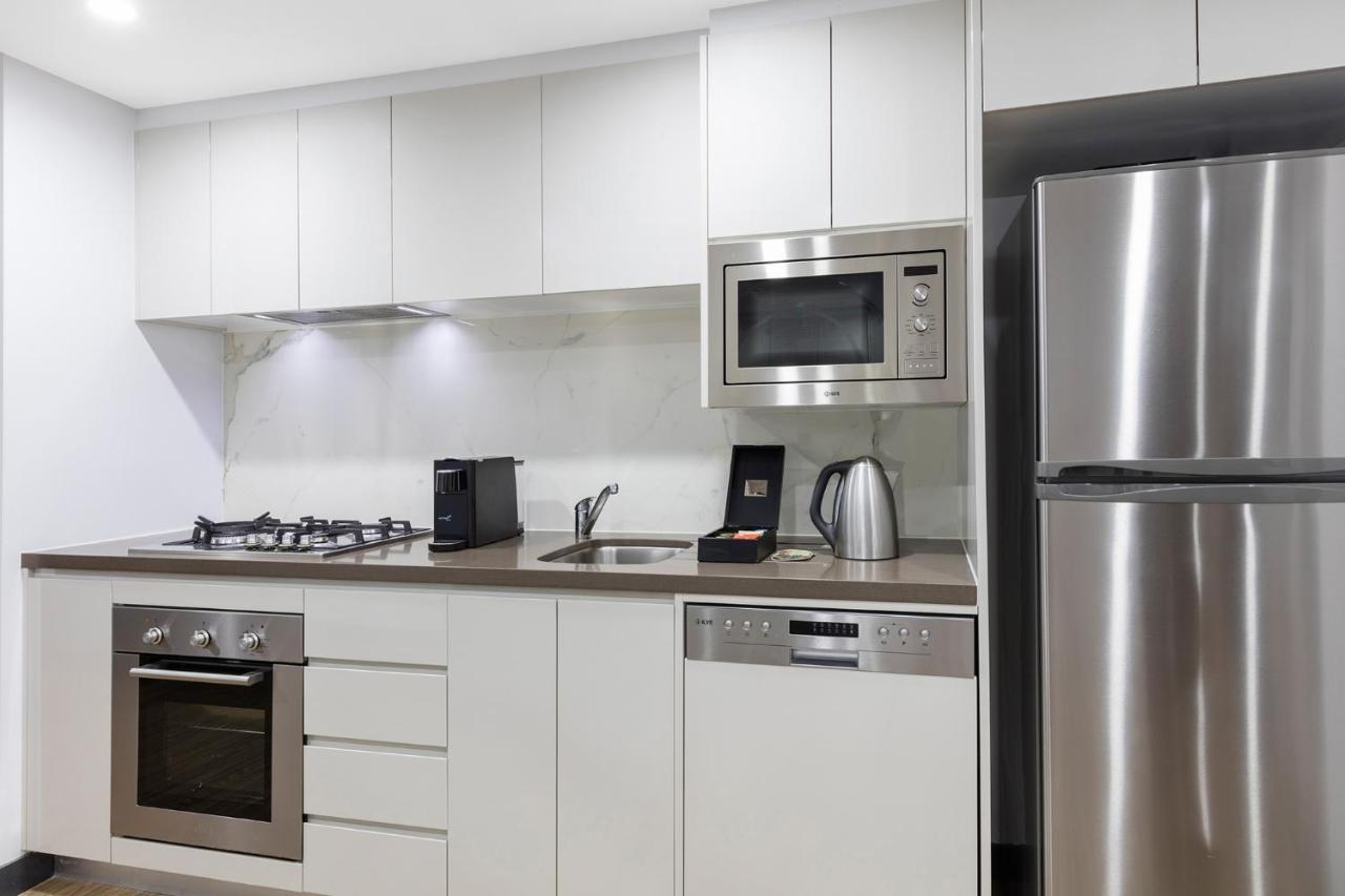 Meriton Suites Bondi Junction Σίδνεϊ Εξωτερικό φωτογραφία The photo shows a modern kitchen with a sleek, minimalist design. It features white cabinetry and a countertop that blends with the overall aesthetic. 

Key elements include:

- A stainless steel refrigerator on the right.
- An oven with a gas cookto