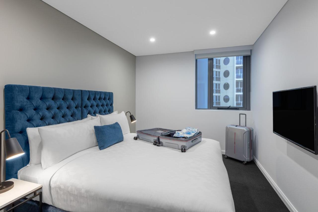 Meriton Suites Bondi Junction Σίδνεϊ Εξωτερικό φωτογραφία The photo shows a modern hotel room featuring a neatly made bed with a blue tufted headboard. There is a small blue decorative pillow on the bed, and the bedding is white. Next to the bed, there is a nightstand with a lamp. A suitcase is partially op