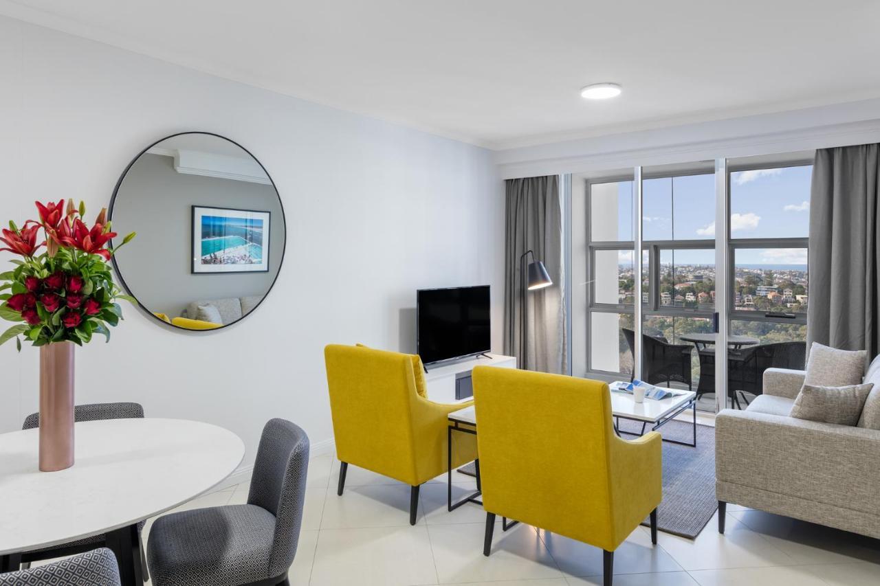 Meriton Suites Bondi Junction Σίδνεϊ Εξωτερικό φωτογραφία The photo shows a modern living space with a stylish design. There are two bright yellow chairs facing a small glass table, and a comfortable beige sofa to the right. A TV is positioned on the wall in front of the chairs. To the left, there's a dinin