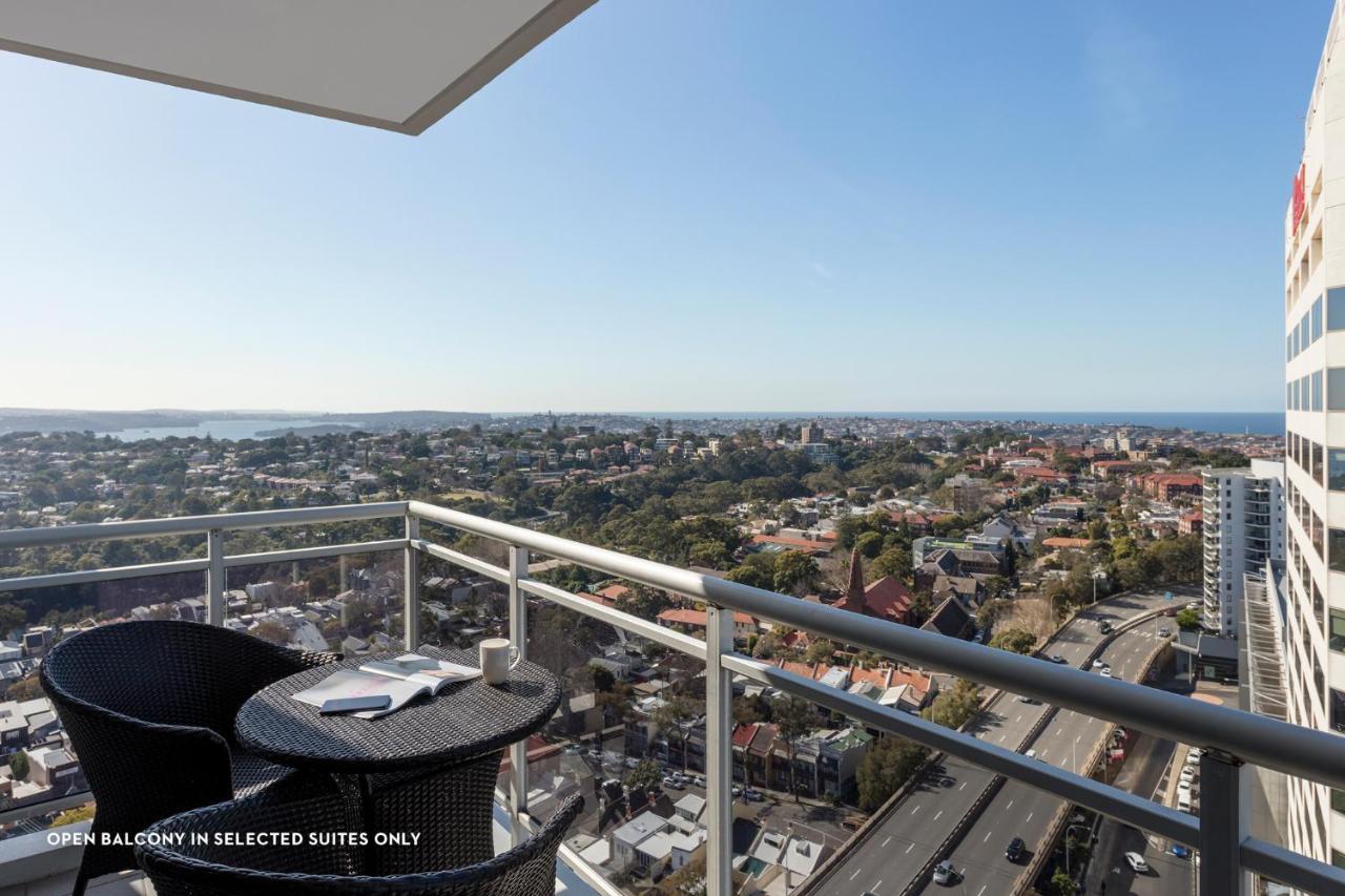 Meriton Suites Bondi Junction Σίδνεϊ Εξωτερικό φωτογραφία The photo shows a balcony with a view over a cityscape that includes buildings and greenery, extending towards the horizon where the sea is visible. The balcony features a small round table and two black chairs, suggesting a space for relaxation or d