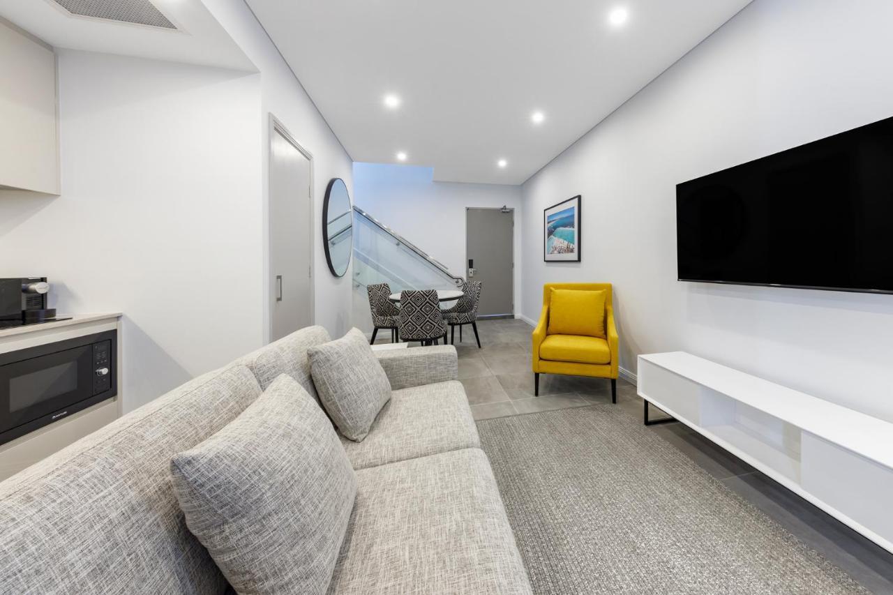 Meriton Suites Bondi Junction Σίδνεϊ Εξωτερικό φωτογραφία The photo shows a modern and stylish living space. It features a light-colored sofa with cushions, positioned to face a wall-mounted flat-screen TV. There's a yellow accent chair nearby that adds a pop of color to the room. The décor is minimalistic,