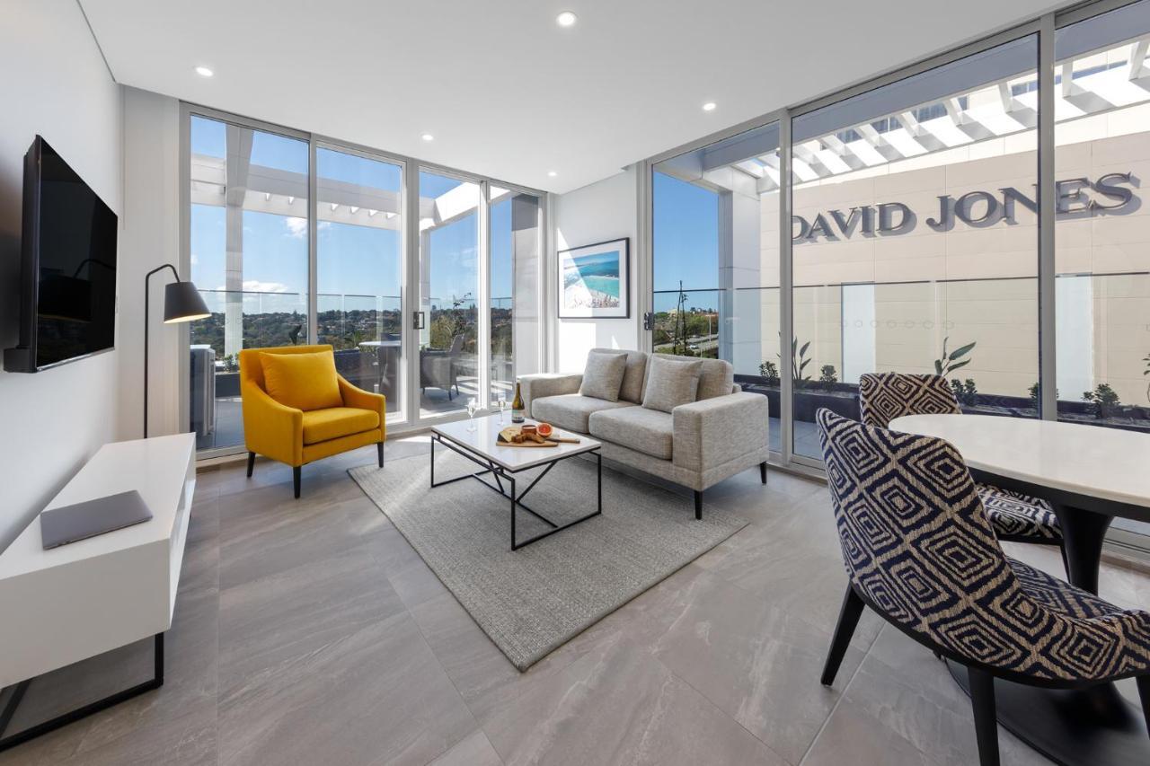 Meriton Suites Bondi Junction Σίδνεϊ Εξωτερικό φωτογραφία The photo shows a modern living space that features large windows allowing ample natural light. The room is furnished with a gray sofa and a bright yellow armchair, along with a coffee table in between. There’s a decorative bowl with fruits on the ta