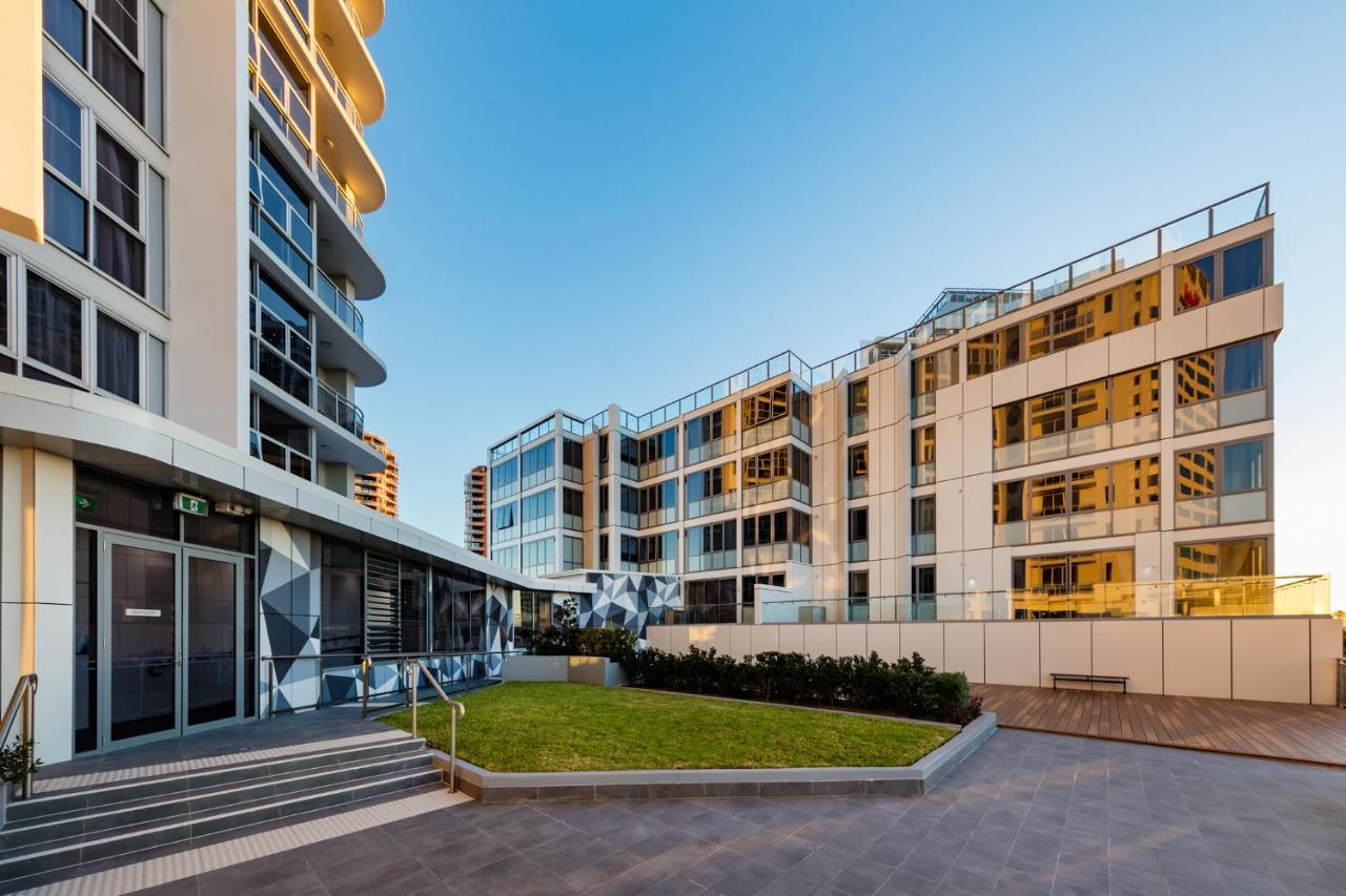 Meriton Suites Bondi Junction Σίδνεϊ Εξωτερικό φωτογραφία The photo showcases a modern residential area featuring contemporary buildings. In the foreground, there is a landscaped green space surrounded by well-designed architecture. The buildings have large glass windows, reflecting the light, and feature a