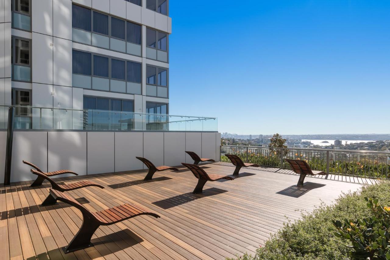 Meriton Suites Bondi Junction Σίδνεϊ Εξωτερικό φωτογραφία The photo shows a rooftop or outdoor area with a wooden deck. There are several modern lounge chairs arranged neatly, facing a clear sky and a panoramic view of the surrounding landscape. In the background, there is a tall building with large windows