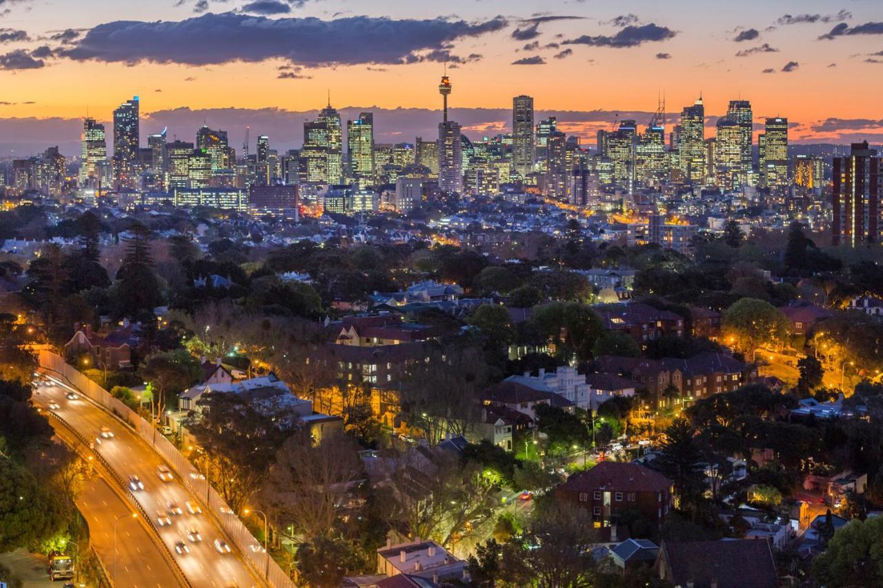 Meriton Suites Bondi Junction Σίδνεϊ Εξωτερικό φωτογραφία The photo captures a stunning city skyline at dusk, featuring towering skyscrapers adorned with lights against a colorful sunset backdrop. In the foreground, a green area with trees and houses is visible, while a road with cars can be seen winding th