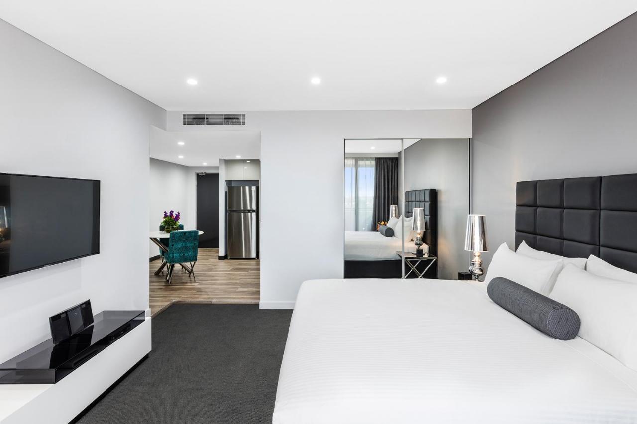 Meriton Suites Bondi Junction Σίδνεϊ Εξωτερικό φωτογραφία The photo shows a modern hotel room. It features a large bed with a white covering and gray accent pillows. To the left, there's a flat-screen TV mounted on the wall above a sleek white cabinet. The flooring is a light wood tone, and a large mirror i