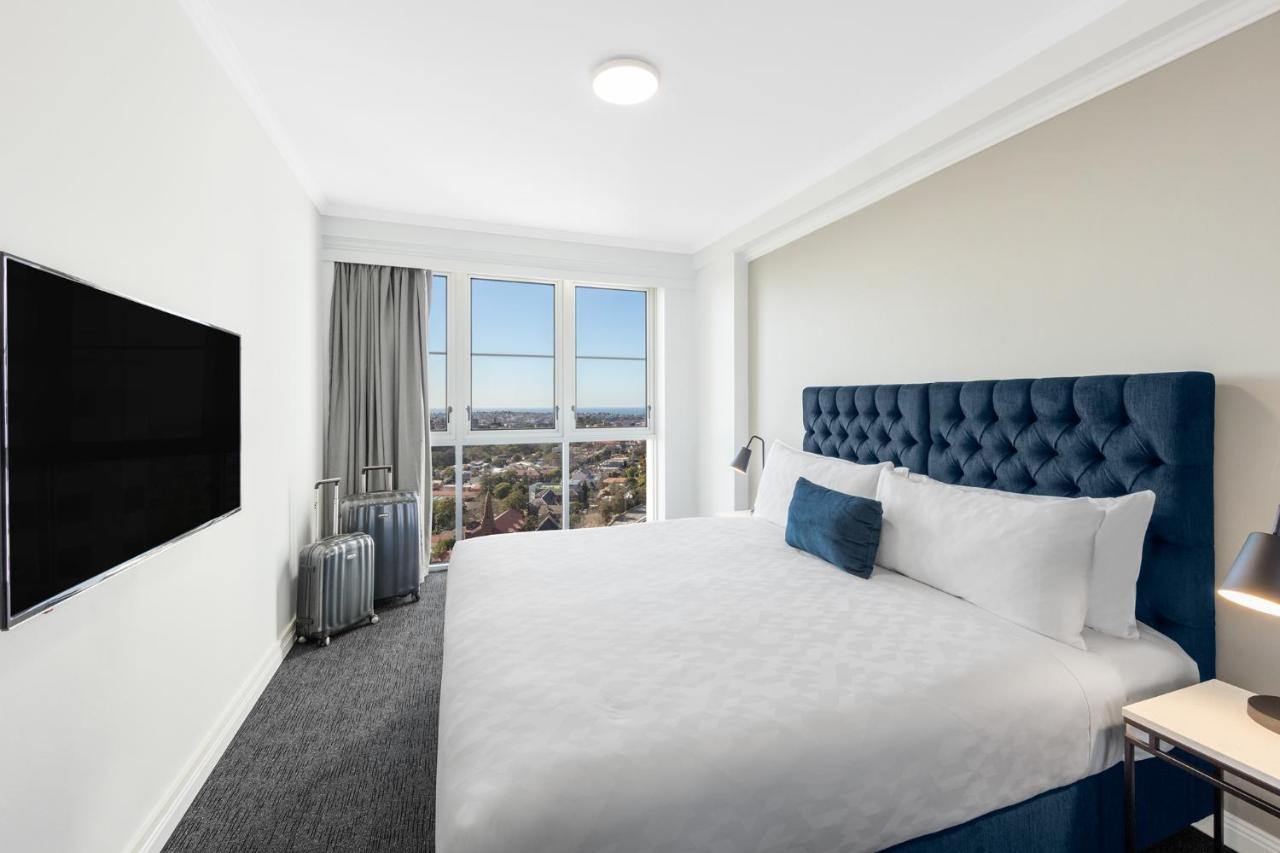 Meriton Suites Bondi Junction Σίδνεϊ Εξωτερικό φωτογραφία The photo shows a modern hotel room with a large bed featuring a tufted blue headboard. There is a white duvet and a decorative pillow on the bed. A flat-screen television is mounted on the wall opposite the bed. Large windows are covered with gray c