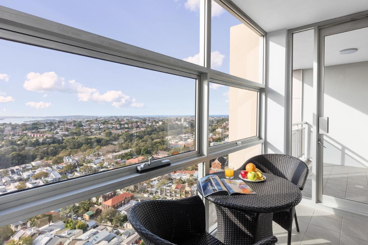 Meriton Suites Bondi Junction Σίδνεϊ Εξωτερικό φωτογραφία The photo shows a modern balcony with large glass windows that offer a panoramic view of a sprawling urban landscape. There is a small round table made of dark wicker, accompanied by two matching chairs. On the table, there is a bowl of fruit and a m
