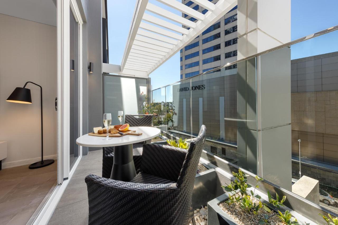 Meriton Suites Bondi Junction Σίδνεϊ Εξωτερικό φωτογραφία The photo shows a modern balcony area with glass railings. It features a small round table set for two, with a plate of food and drinks. There are two stylish wicker chairs positioned around the table. The space is bright and airy, with sunlight stre