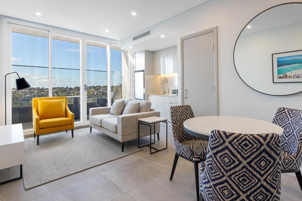 Meriton Suites Bondi Junction Σίδνεϊ Εξωτερικό φωτογραφία The photo shows a modern and stylish living area. It features a comfortable sofa in a neutral color, accompanied by a bright yellow armchair that adds a pop of color. There's a round dining table with patterned chairs nearby. Large windows allow plen