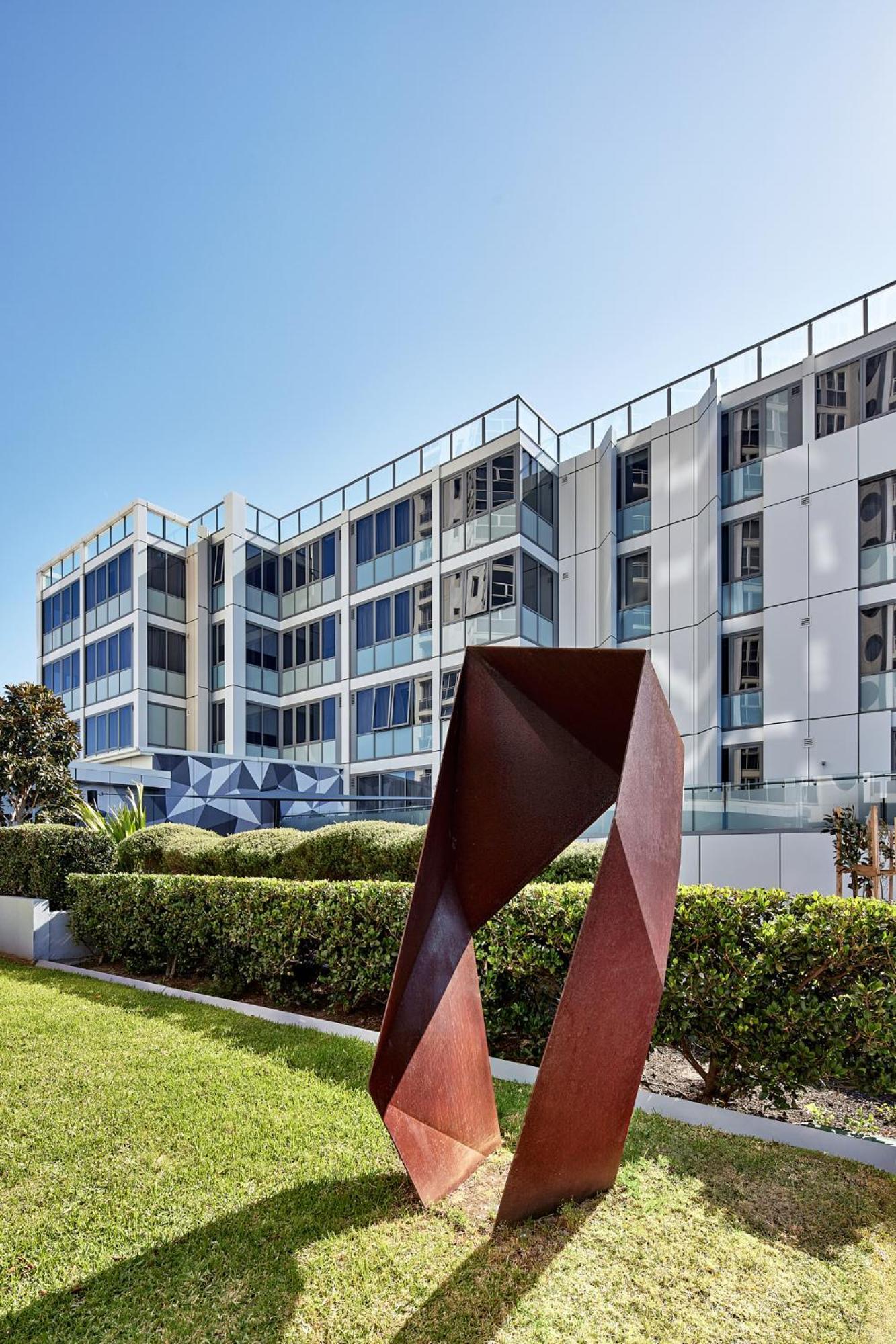 Meriton Suites Bondi Junction Σίδνεϊ Εξωτερικό φωτογραφία The photo shows a modern building with a sleek white and glass facade, featuring multiple floors and large windows. In the foreground, there is a sculptural piece made of metal, characterized by geometric shapes and a reddish-brown color. The surroun