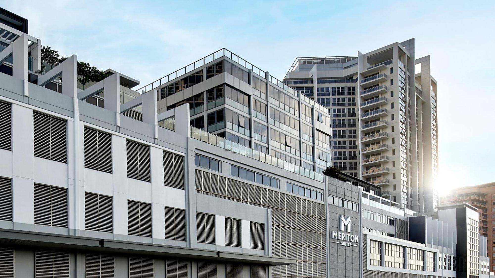 Meriton Suites Bondi Junction Σίδνεϊ Εξωτερικό φωτογραφία The image shows a modern urban building or complex. The structure features a combination of glass and concrete elements, showcasing contemporary architectural design. The foreground has a lower section that appears to be a podium or base with a textu