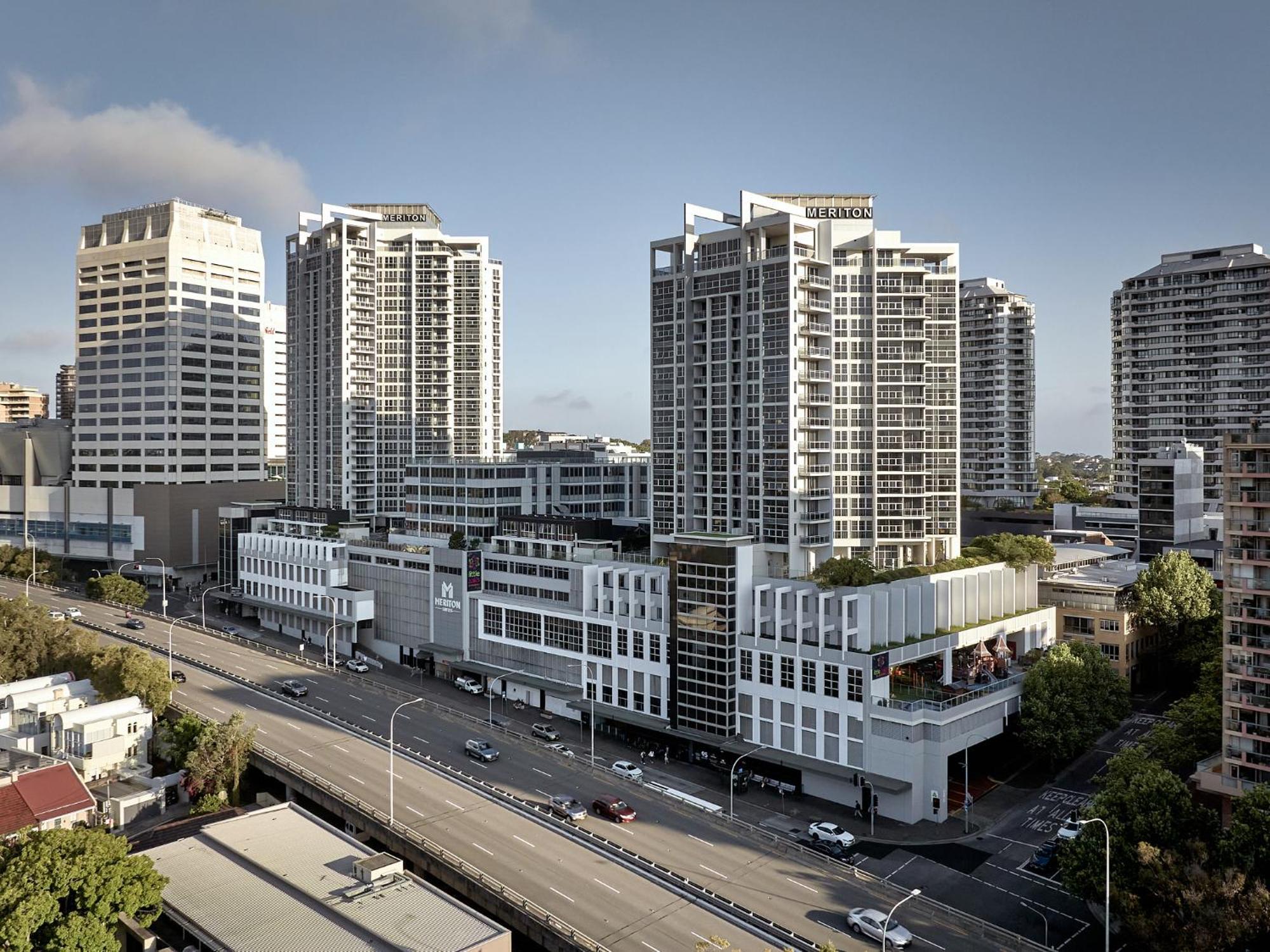 Meriton Suites Bondi Junction Σίδνεϊ Εξωτερικό φωτογραφία The photo shows a cityscape featuring several tall modern buildings, likely apartments or office complexes. In the foreground, there is a road lined with cars, and the architecture of the buildings varies, with some having sleek glass facades and oth