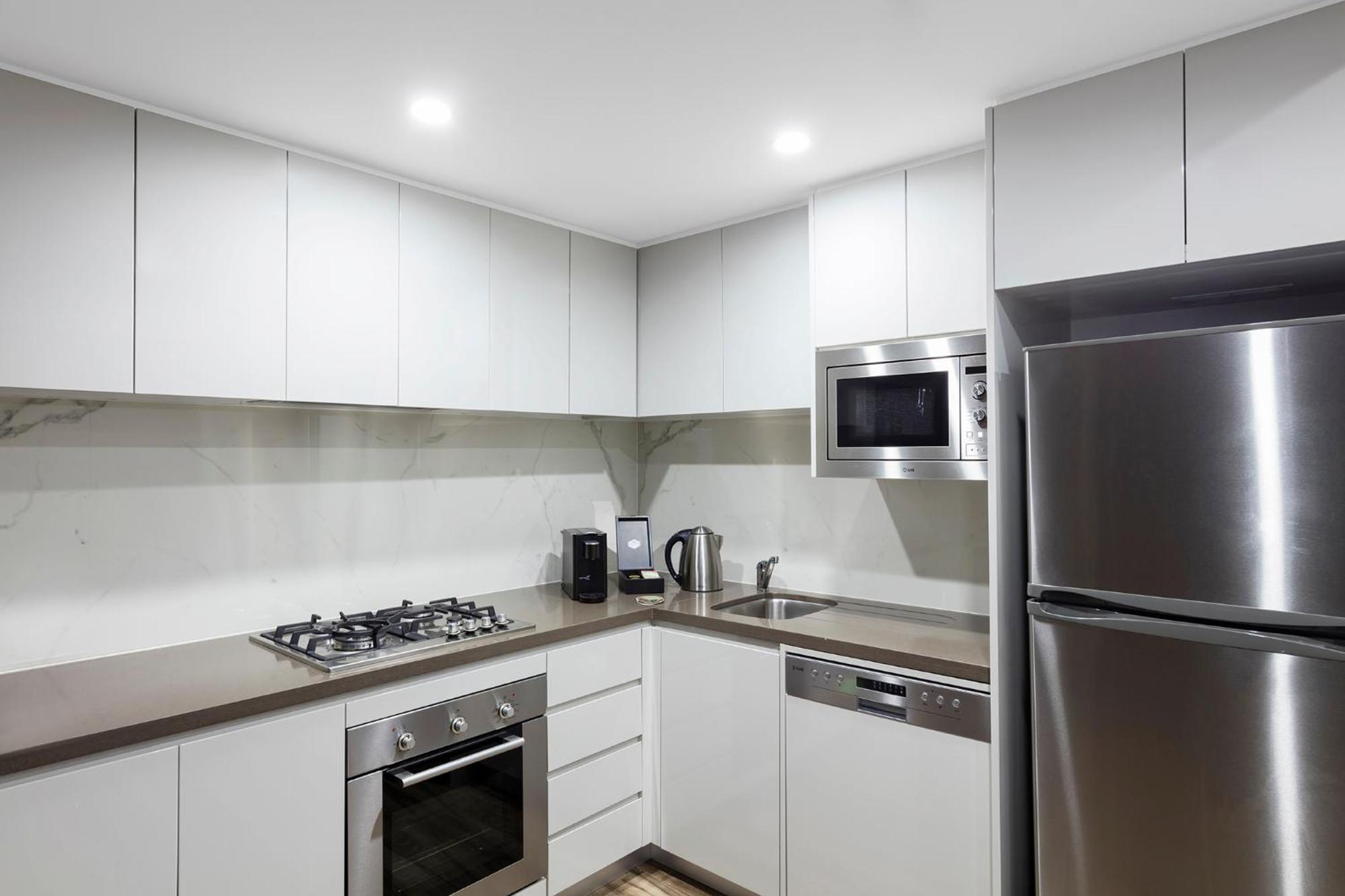 Meriton Suites Bondi Junction Σίδνεϊ Εξωτερικό φωτογραφία The photo shows a modern kitchen featuring a minimalist design. It has light-colored cabinetry, a countertop, and stainless steel appliances. The appliances include a gas stove with multiple burners, an oven, a microwave, a kettle, and a refrigerator