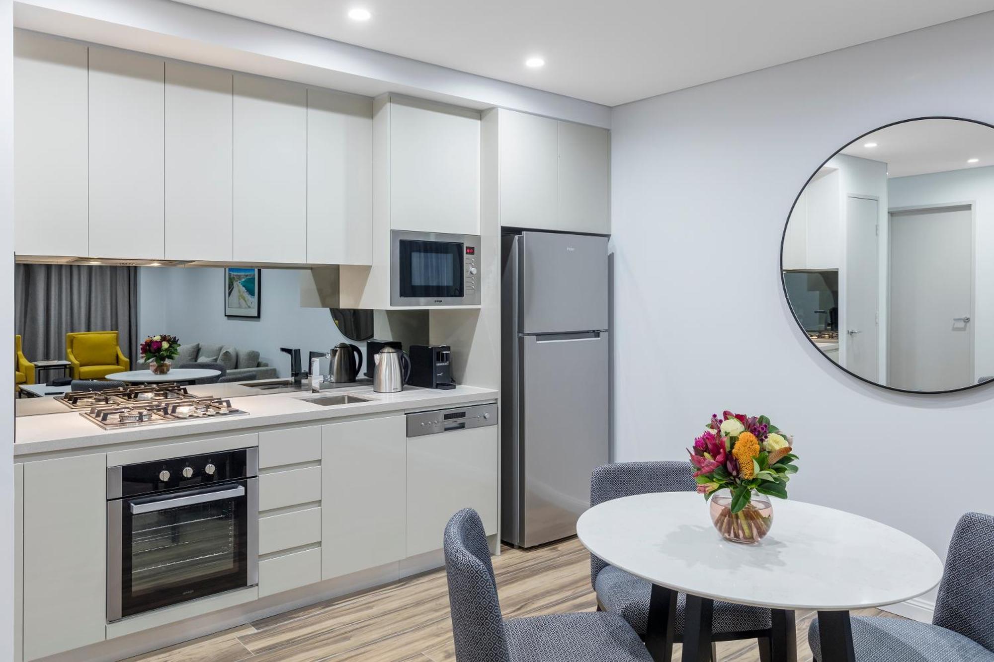 Meriton Suites Bondi Junction Σίδνεϊ Εξωτερικό φωτογραφία The photo shows a modern kitchen and dining area. The kitchen features light-colored cabinetry with a built-in oven and stovetop, a microwave, and a silver refrigerator. There is a kettle and a coffee maker on the countertop. In the dining area, ther
